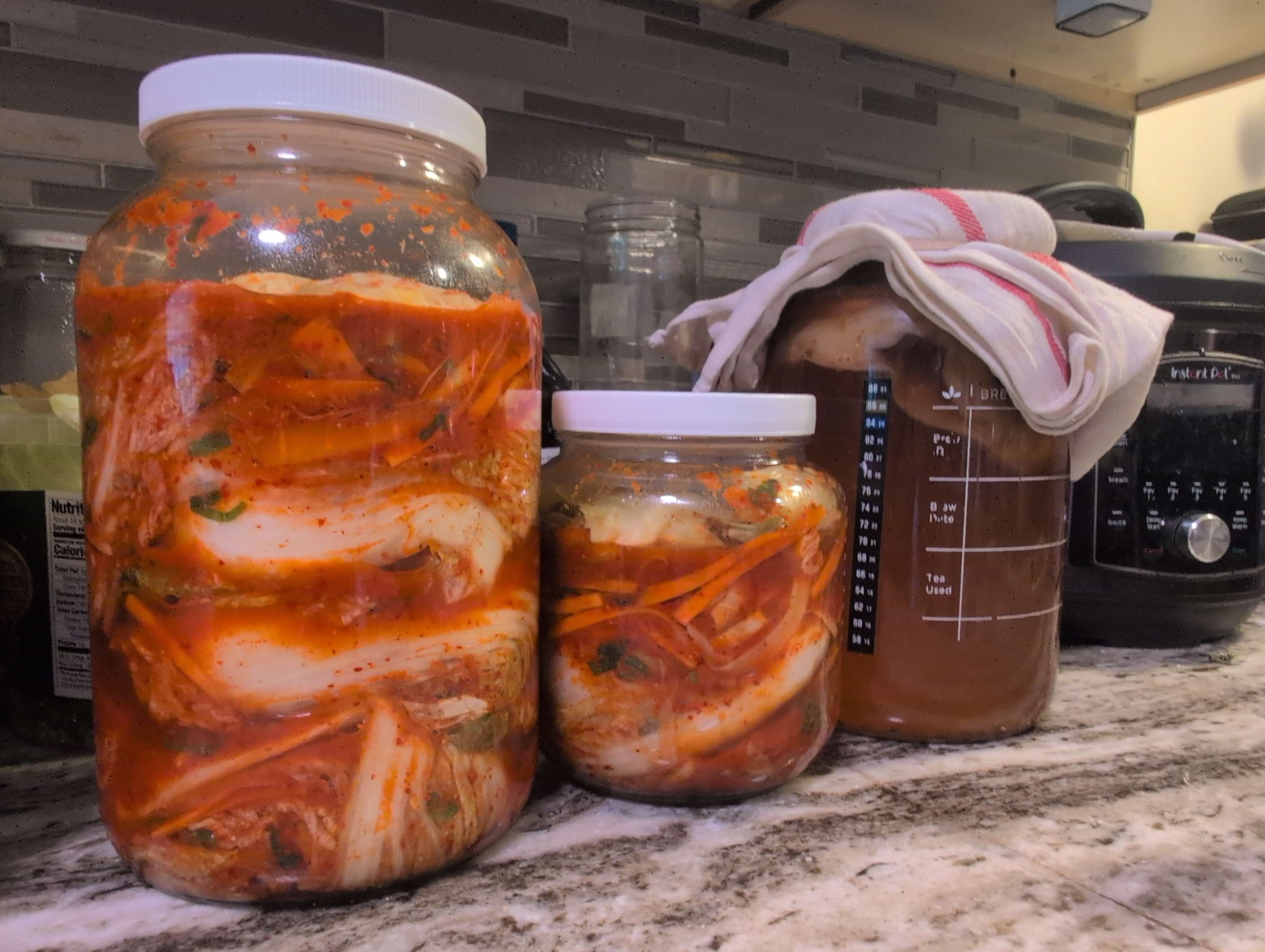 One large gallon jar filled with kimchi that's in the process of fermenting and a smaller one about 1/3rd the size that's also filled with kimchi. A gallon jar of kombucha also sits on the counter.